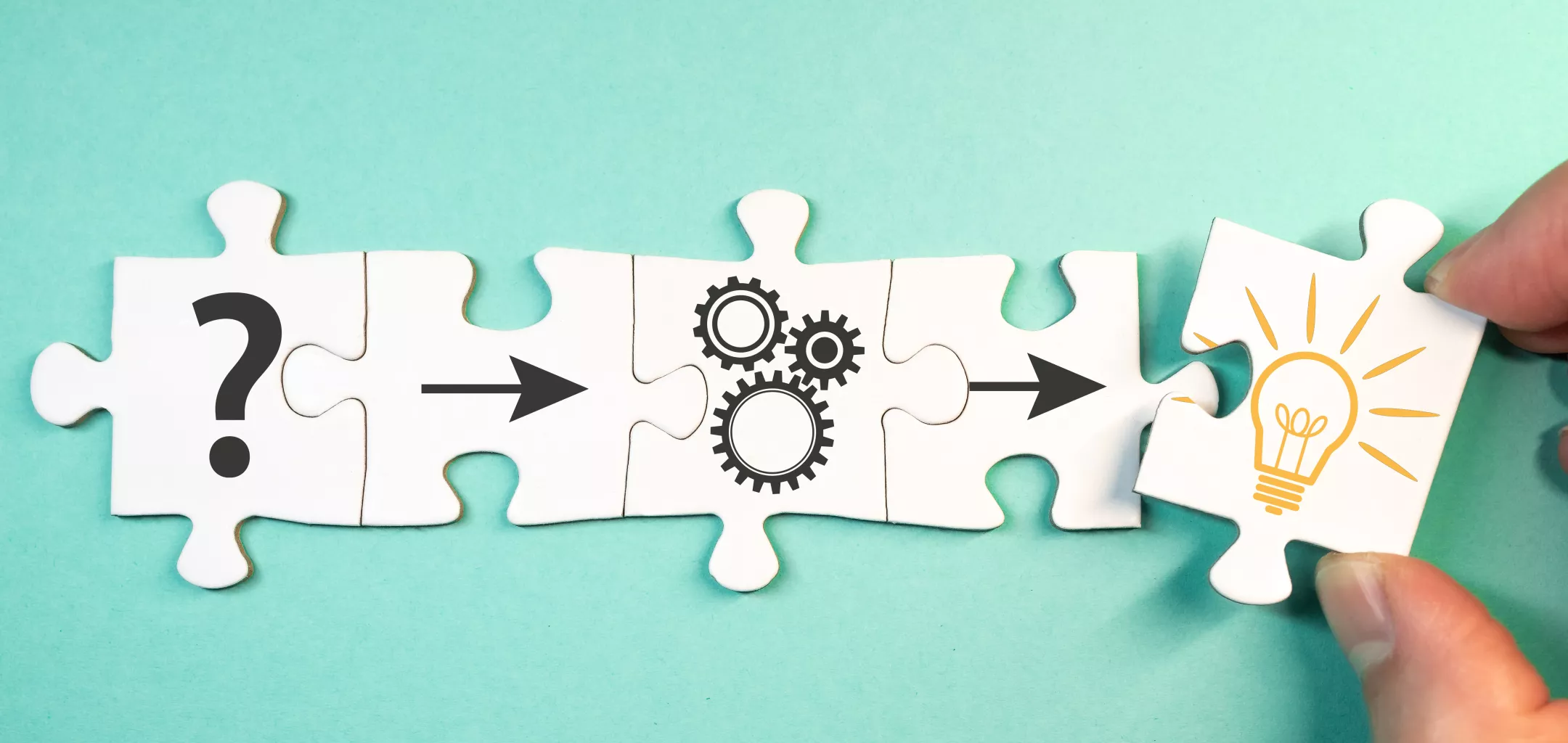 Four puzzle pieces connected in a row on a teal background. The first piece features a question mark, the second an arrow, the third a set of interlocking gears, and the final piece—being placed by a hand—depicts a glowing light bulb. The image symbolizes problem-solving, innovation, or the process of turning questions into solutions.