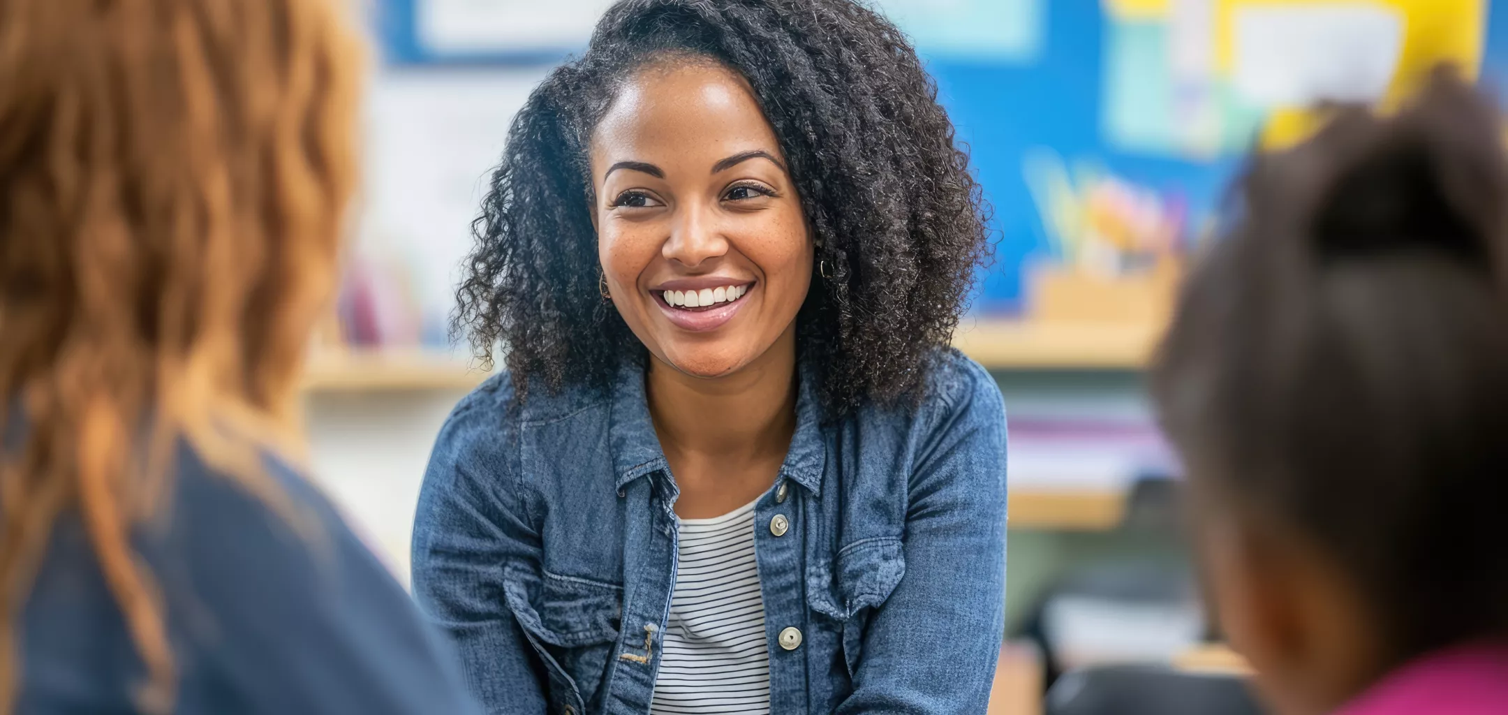A teacher smiling warmly while engaging in a conversation with a parent and a student in a classroom setting. The background shows colorful bulletin boards and educational materials, creating a welcoming and positive learning environment.
