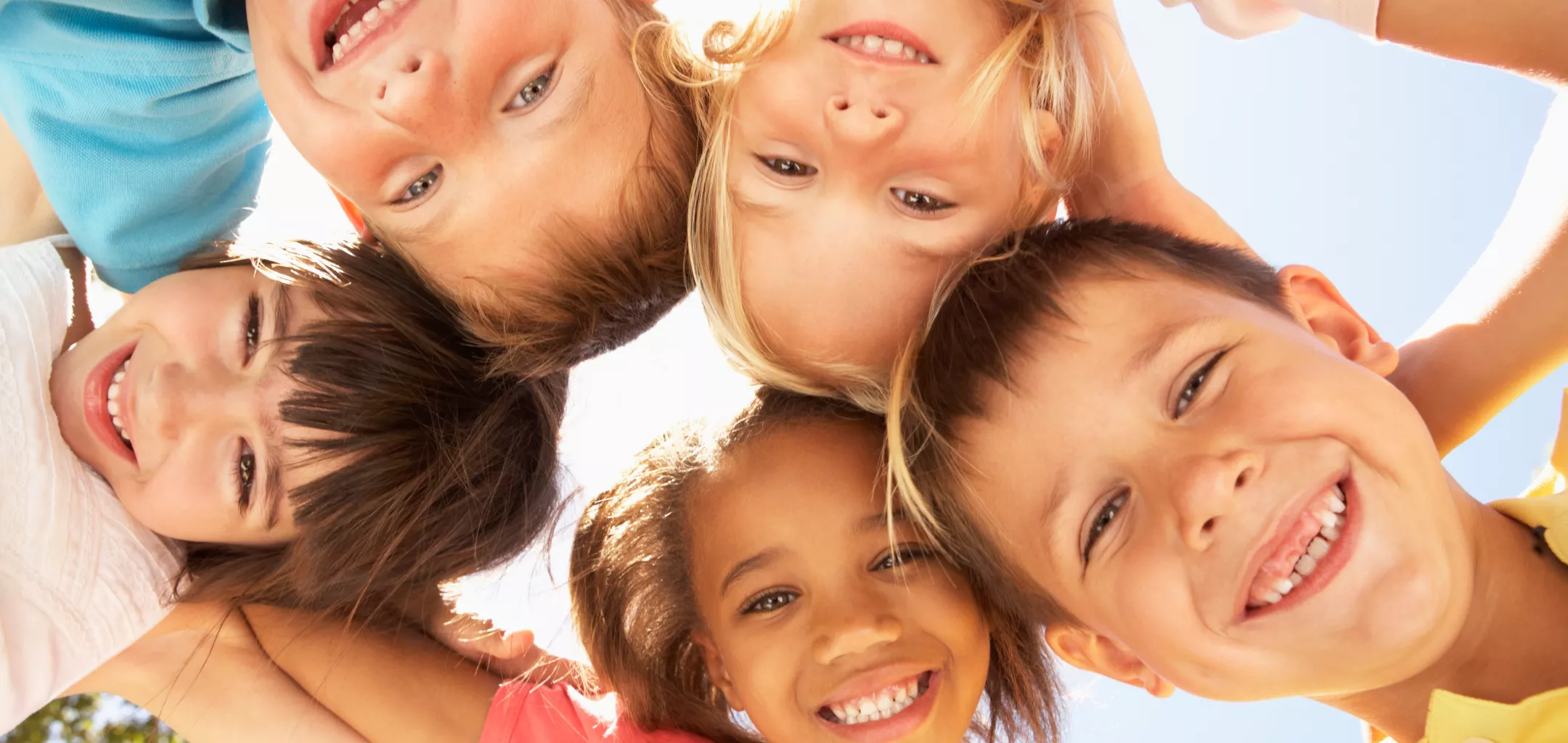A group of five smiling children, heads together in a circle, looking down at the camera against a bright blue sky. The photo is taken from a low angle, capturing their joyful expressions and close friendship.