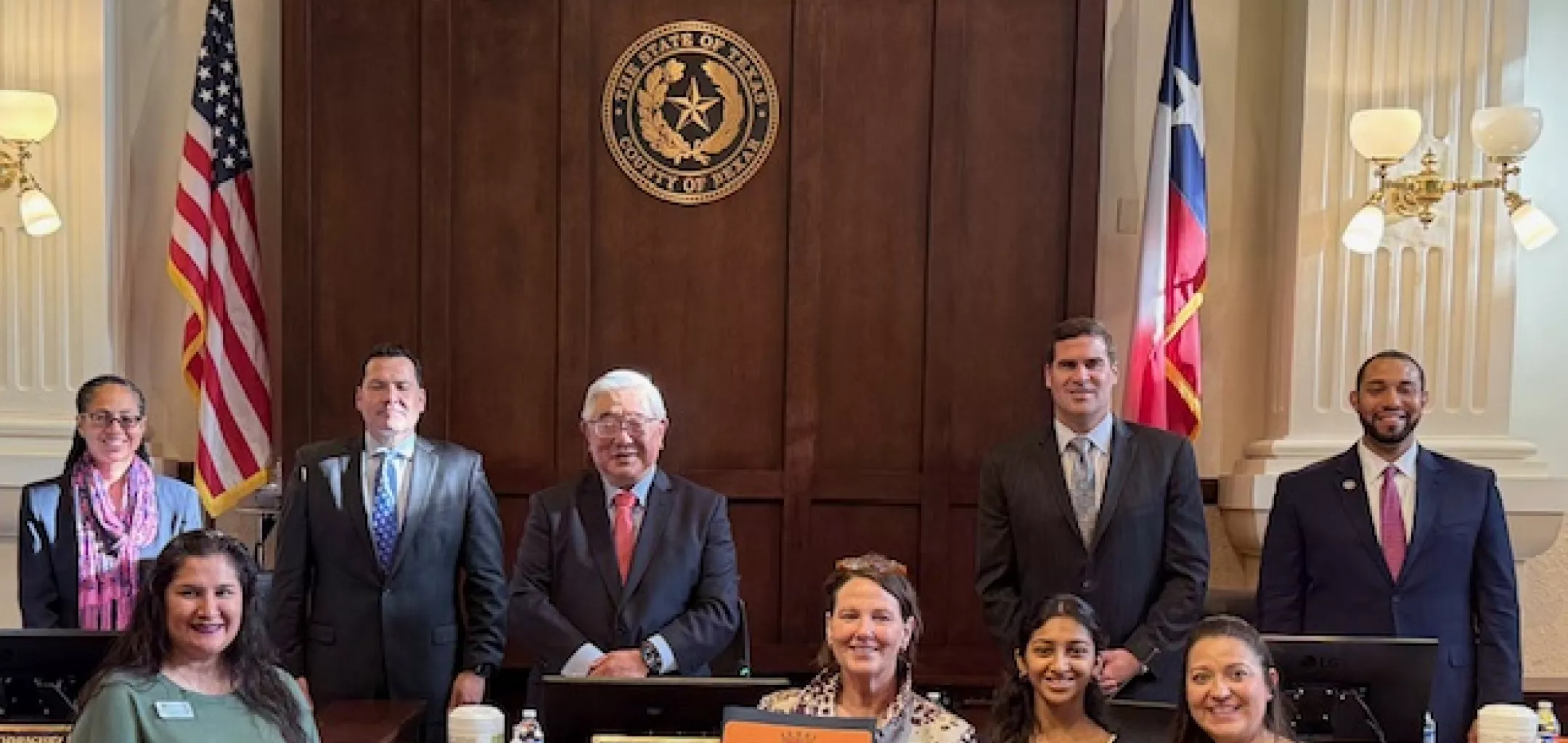 A group of nine individuals stands and smiles inside the Bexar County Courthouse courtroom. Five people, including judges or officials in suits, are standing behind a large wooden bench with a seal of the State of Texas and the words “BEXAR COUNTY COURTHOUSE” above them. In front, four women are standing, one of whom is holding a framed proclamation or certificate. Two American flags and two Texas state flags flank the group. The setting is formal with tall columns, ornate lighting, and wood-paneled decor.